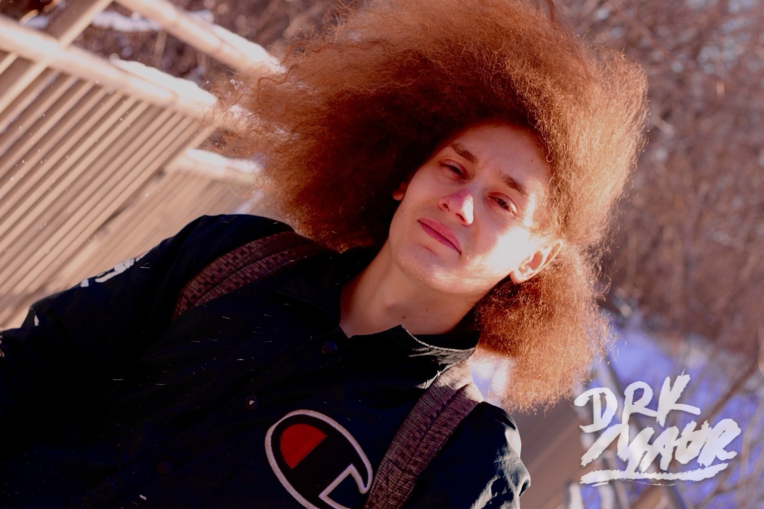 Outdoor portrait of a model with a voluminous reddish-brown afro in a navy Champion jacket against bare winter branches