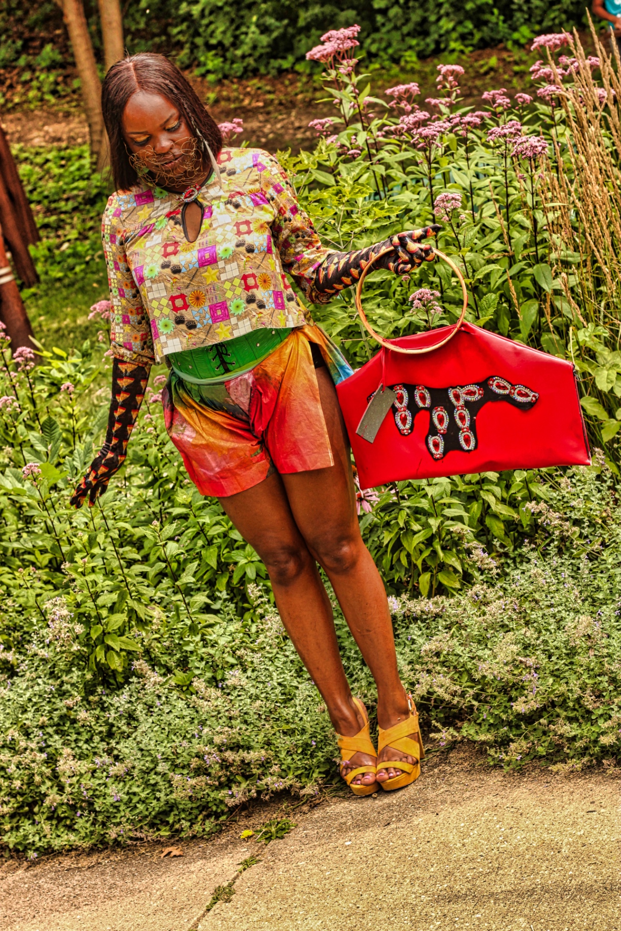 Model in patchwork top and iridescent skirt holding a red structured handbag among tall garden flowers