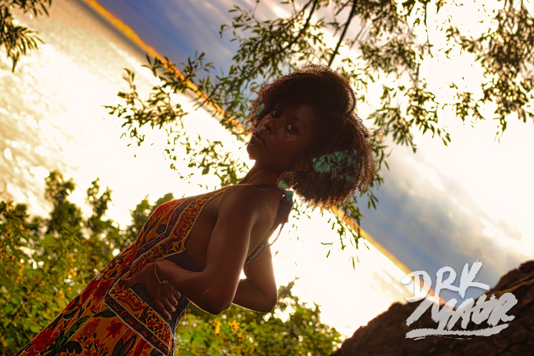 Low-angle portrait of a model in a floral dress leaning into golden backlight through willow branches