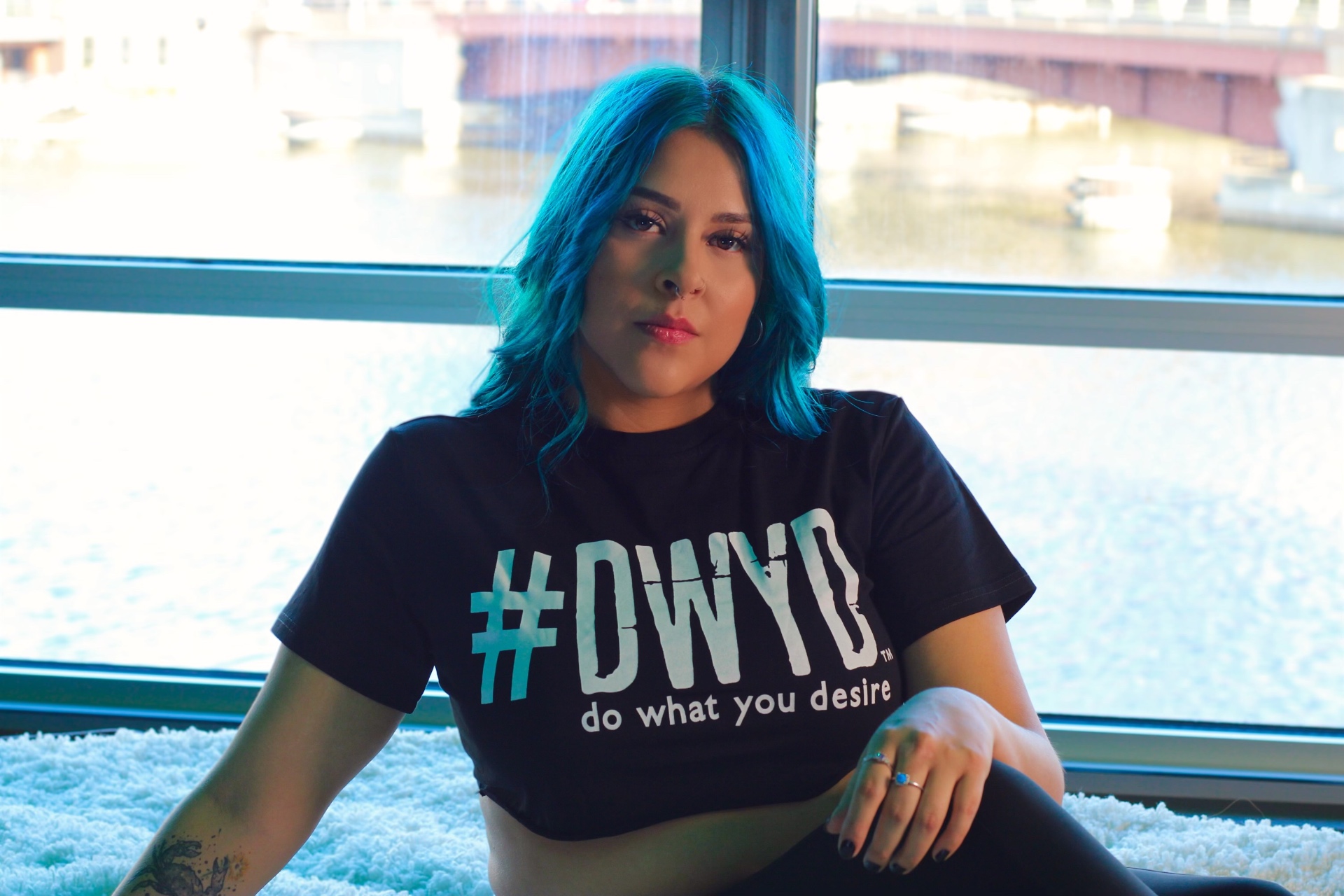 Portrait of a person with blue hair in a graphic tee seated by a bright riverfront window