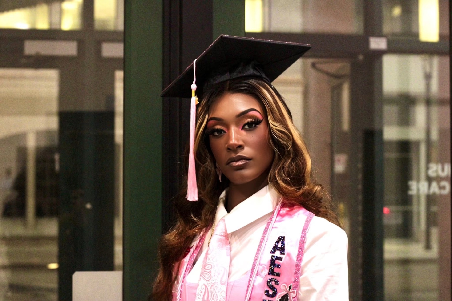 Evening portrait of a graduate in cap and pink esthetician stole against lit storefront glass