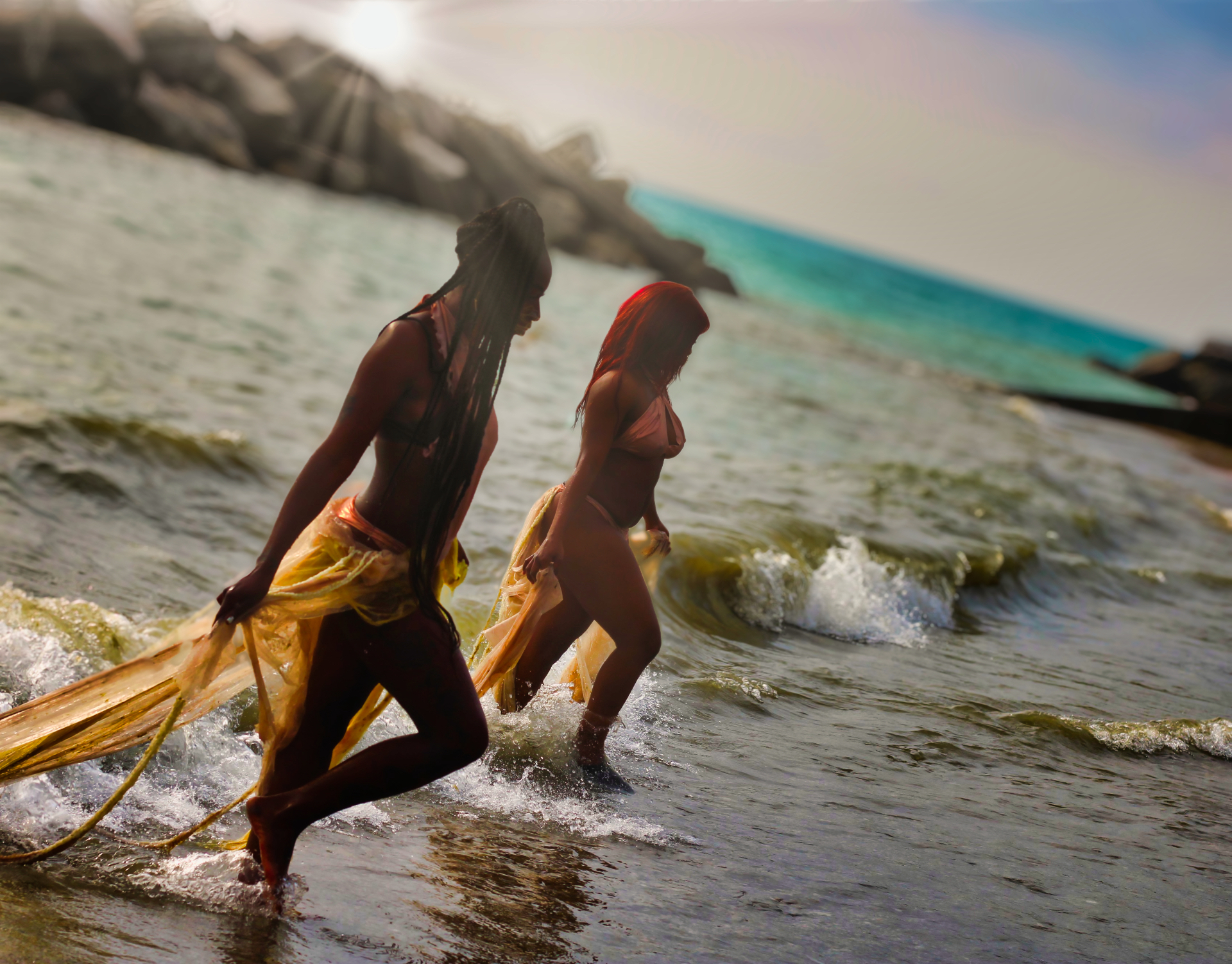 Two models wading through breaking waves trailing gold fabric in backlit sun