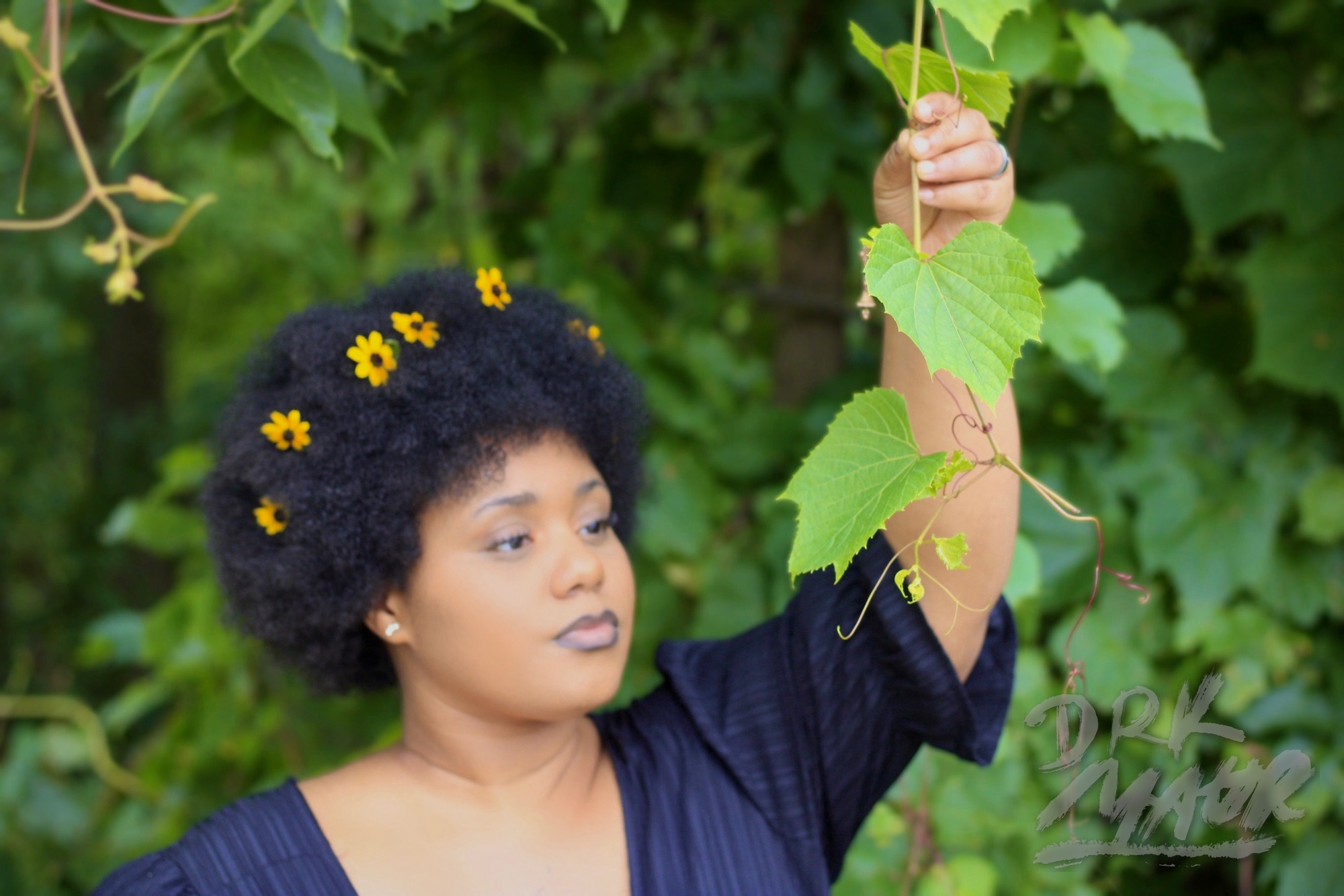Woman in a black dress reaching upward through green grape leaves with yellow flowers in her hair