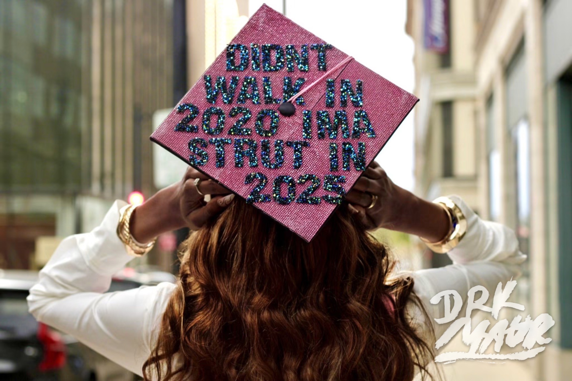 Back view of a graduate holding a pink rhinestone cap reading Didn't walk in 2020 ima strut in 2025