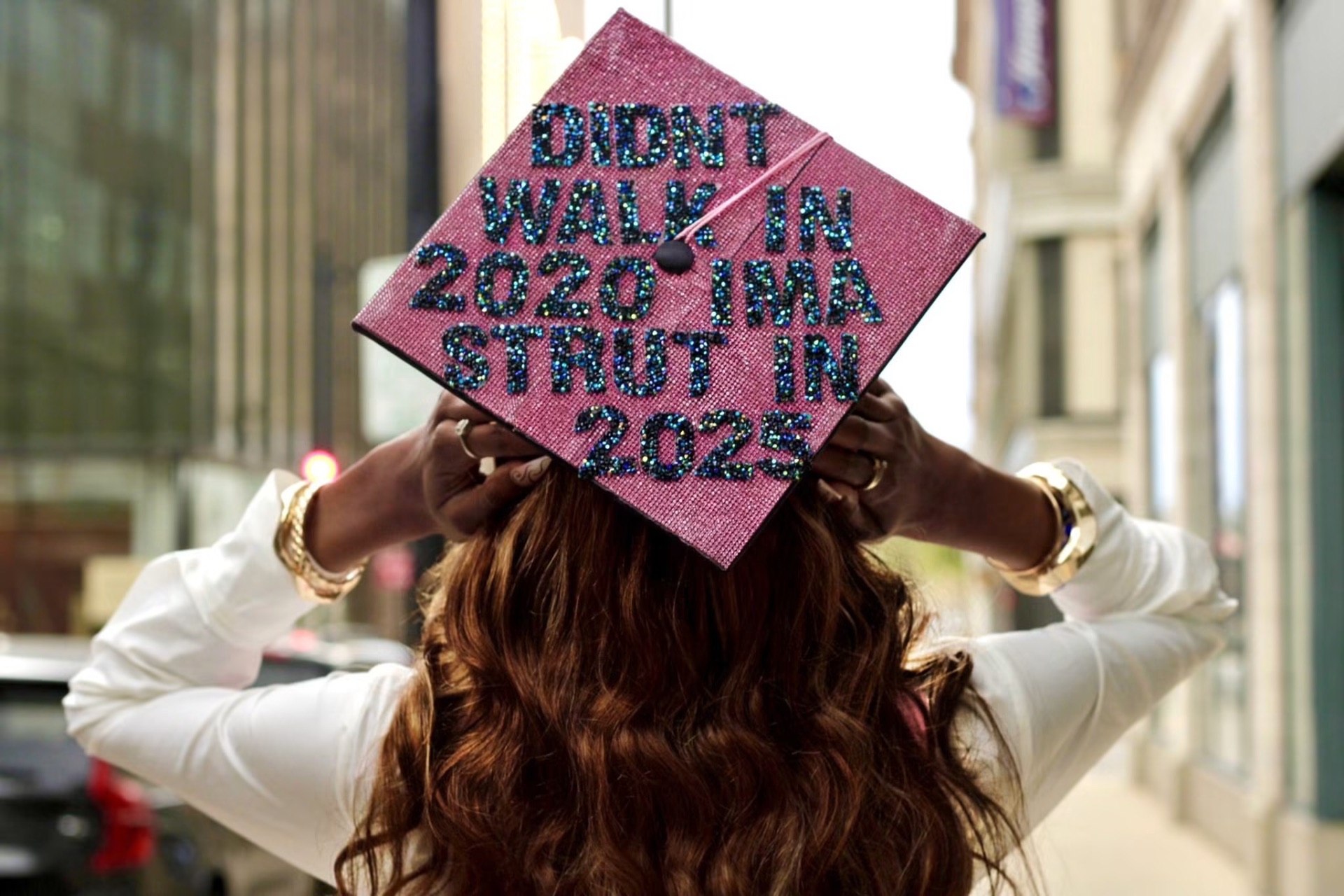 Back view of a graduate holding a pink rhinestone cap reading Didn't walk in 2020 ima strut in 2025