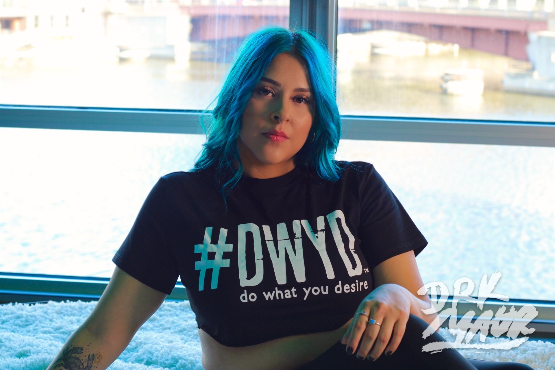 Portrait of a person with blue hair in a graphic tee seated by a bright riverfront window