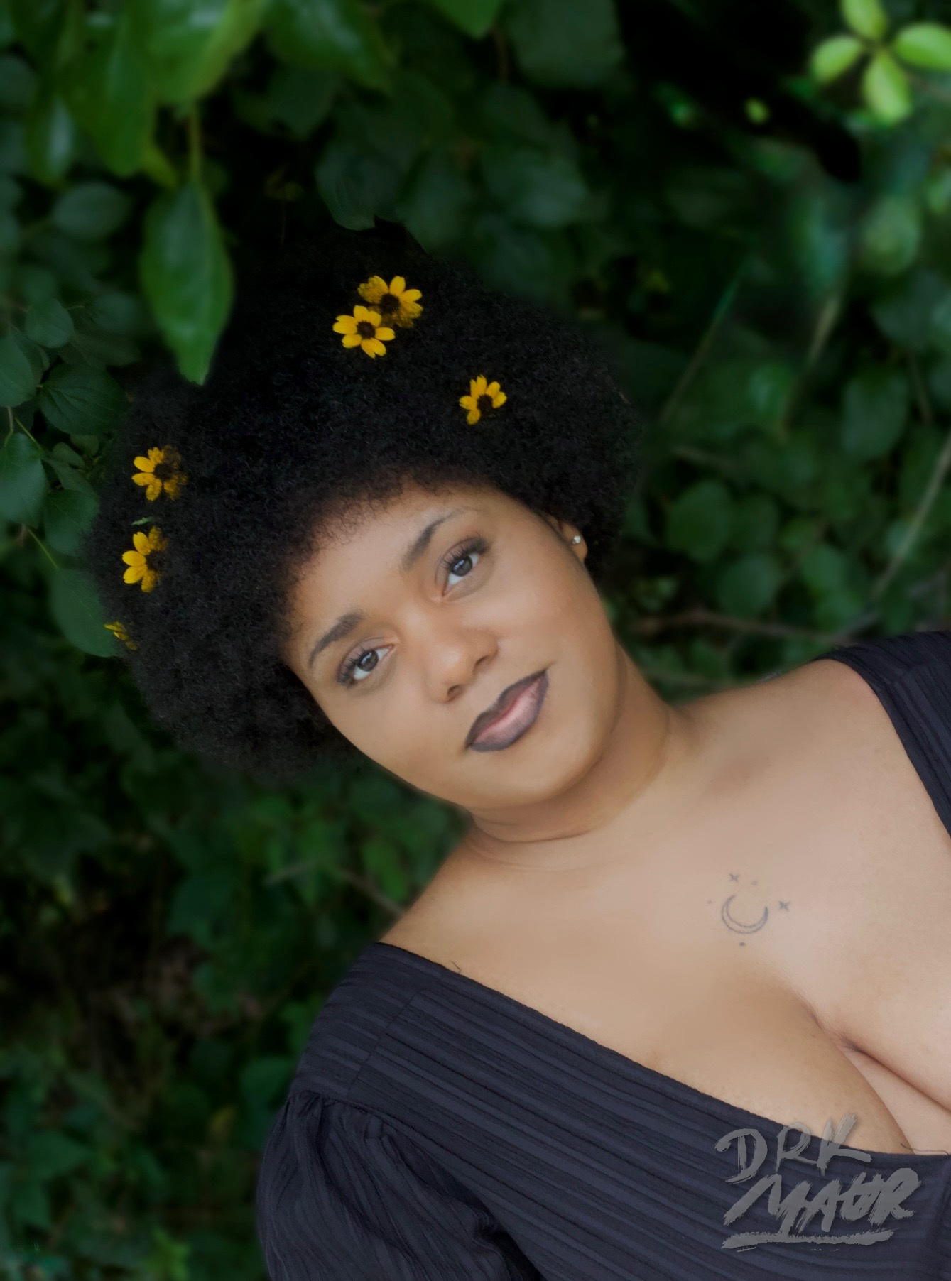 Angled close portrait of a woman in a black dress framed by deep green leaves and yellow flowers