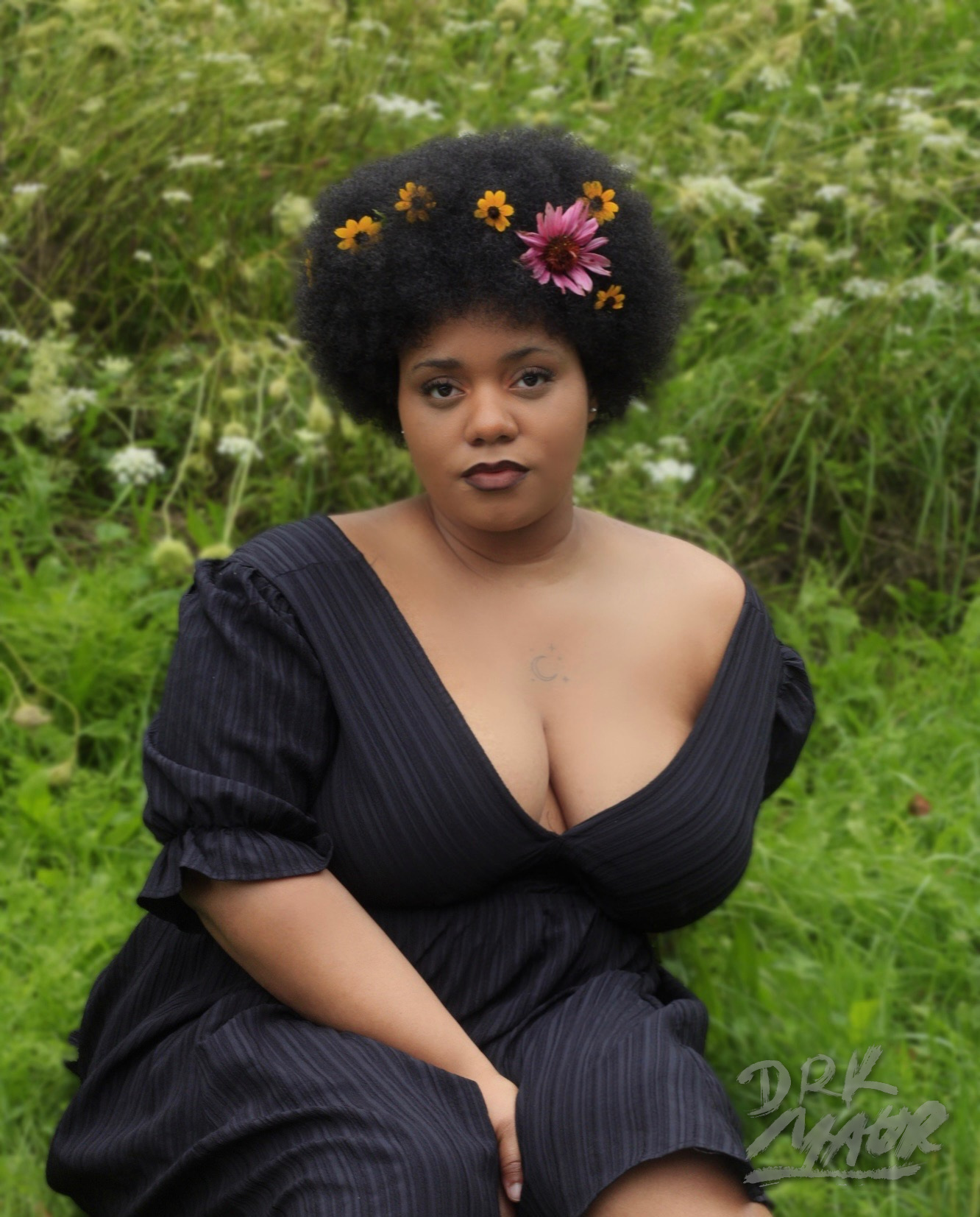 Seated portrait of a woman in a black dress wearing a crown of yellow and purple flowers in a green meadow