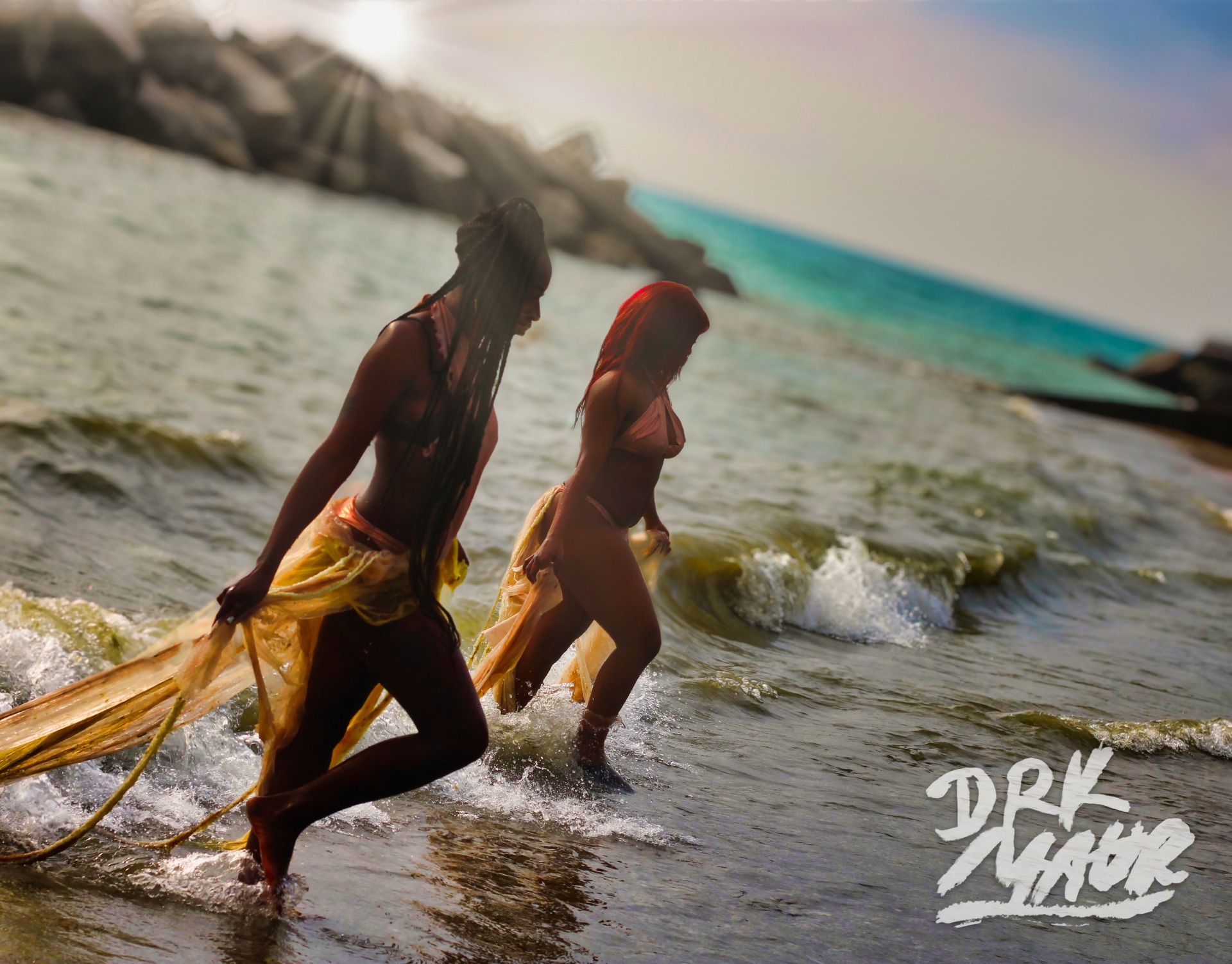 Two models wading through breaking waves trailing gold fabric in backlit sun
