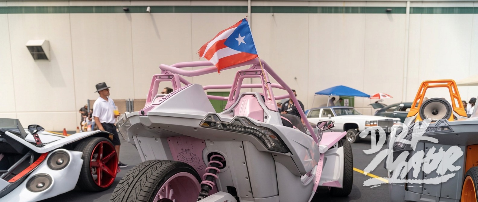 Rear three-quarter view of a pink Polaris Slingshot flying a Puerto Rican flag at a car show