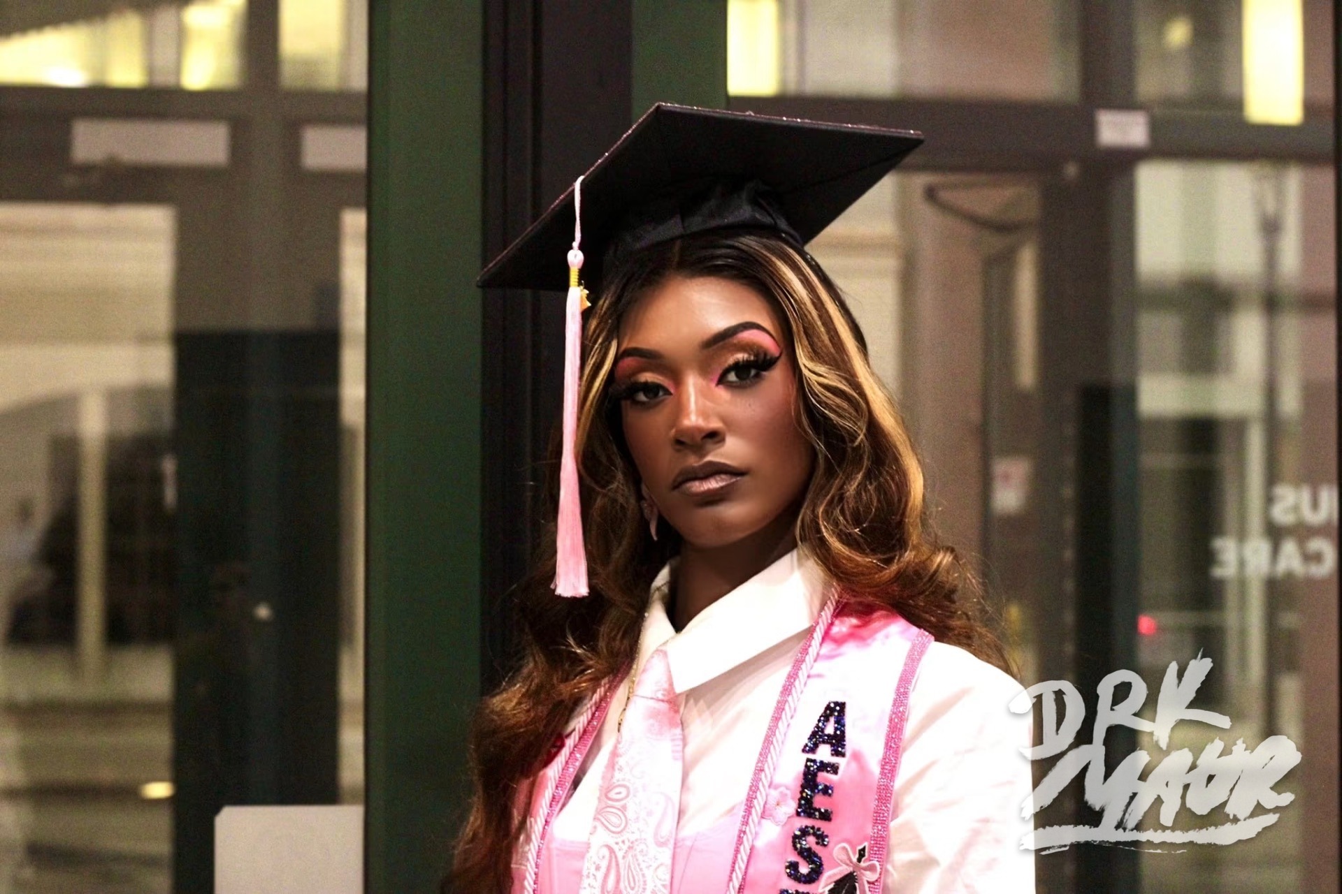 Evening portrait of a graduate in cap and pink esthetician stole against lit storefront glass