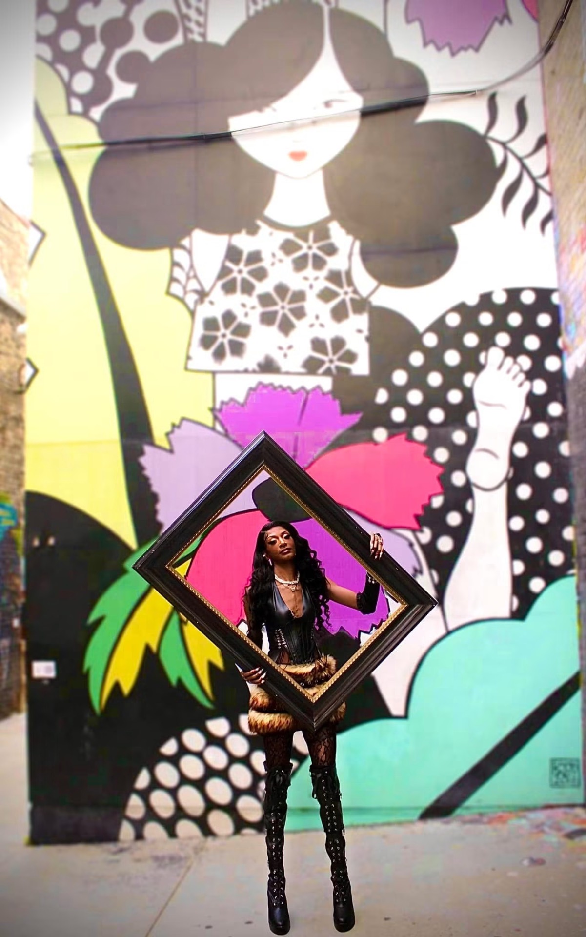 Model holding a tilted gold frame in front of a large pop-art mural