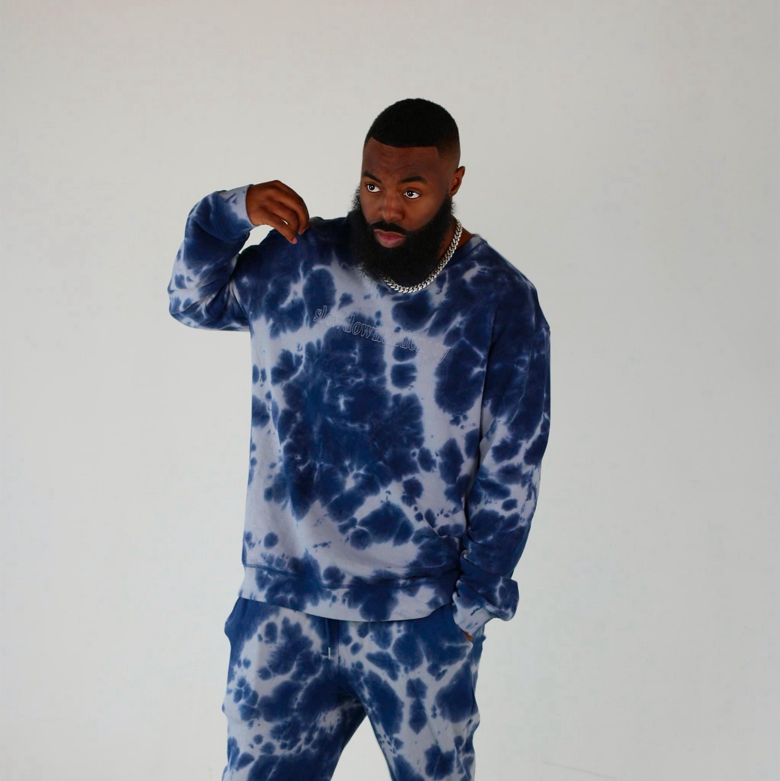 Studio fashion portrait of a bearded model in a navy and steel-blue tie-dye tracksuit embroidered with slow down world