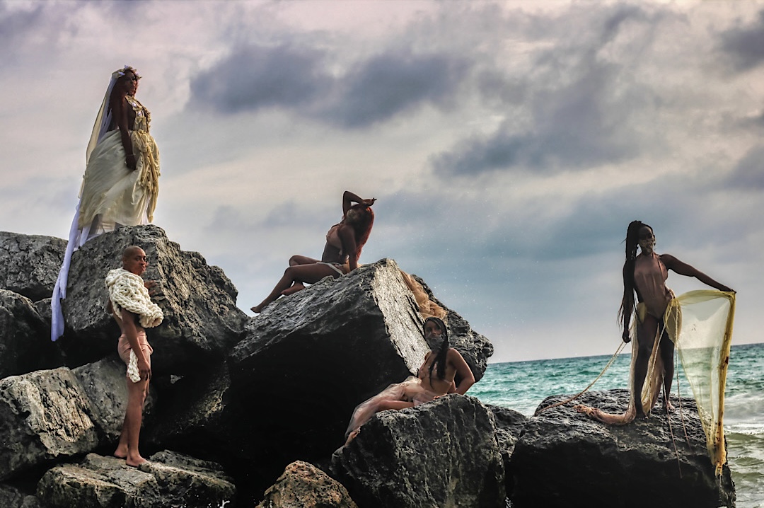 Five models posed across large breakwater rocks beside the water under a dramatic sky
