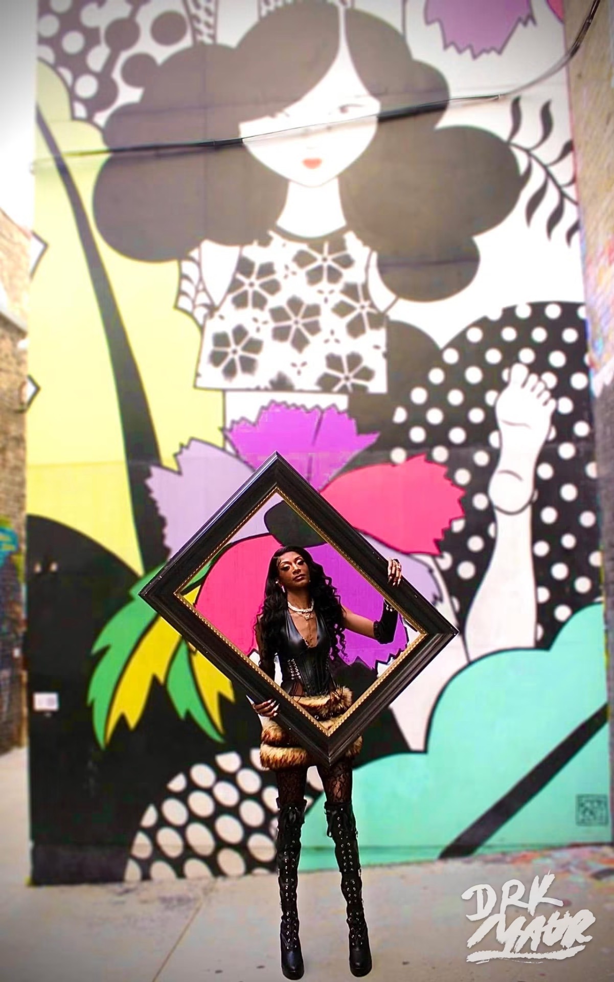 Model holding a tilted gold frame in front of a large pop-art mural