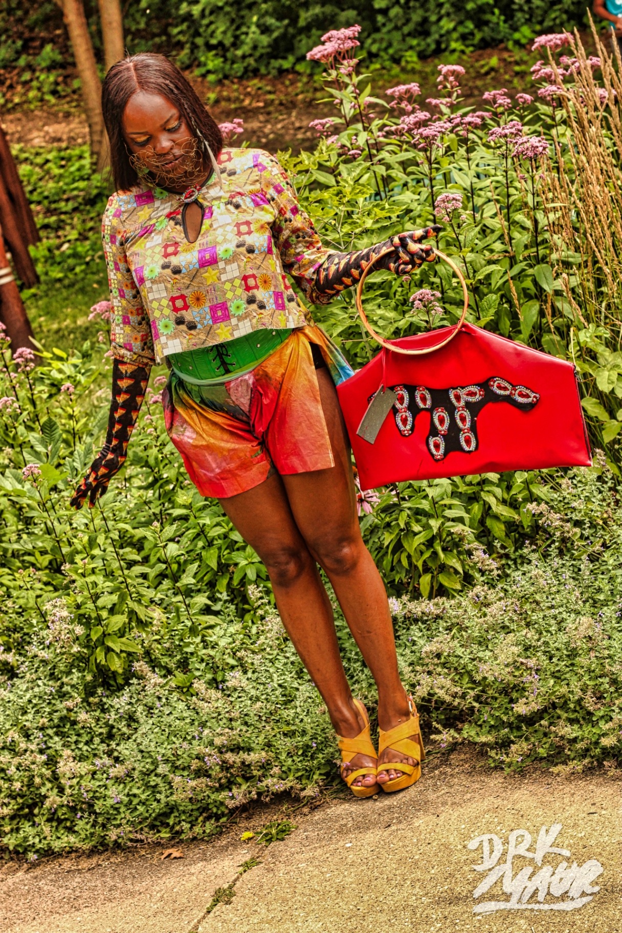 Model in patchwork top and iridescent skirt holding a red structured handbag among tall garden flowers