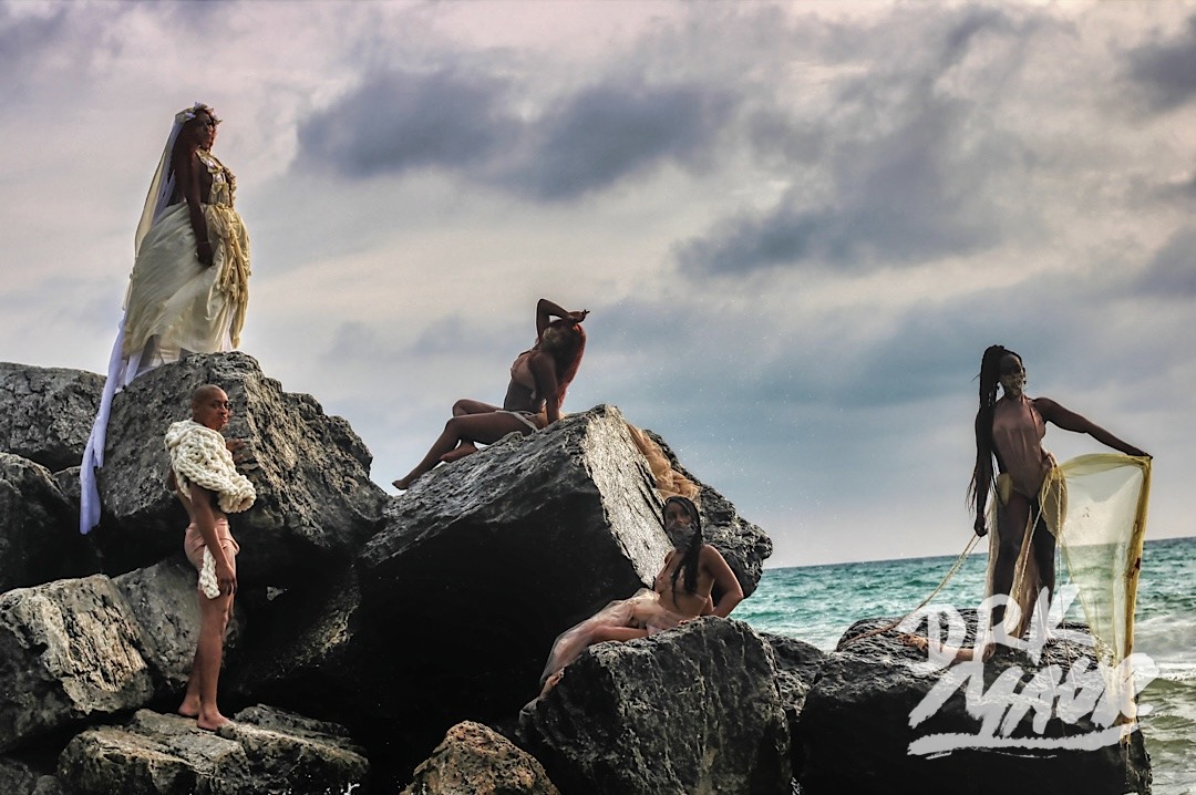 Five models posed across large breakwater rocks beside the water under a dramatic sky