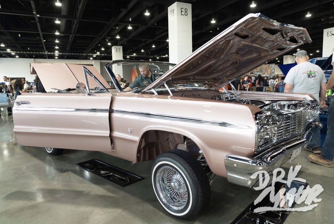 Rose gold Chevrolet Impala convertible lowrider with its hood and trunk open on display at an indoor car show